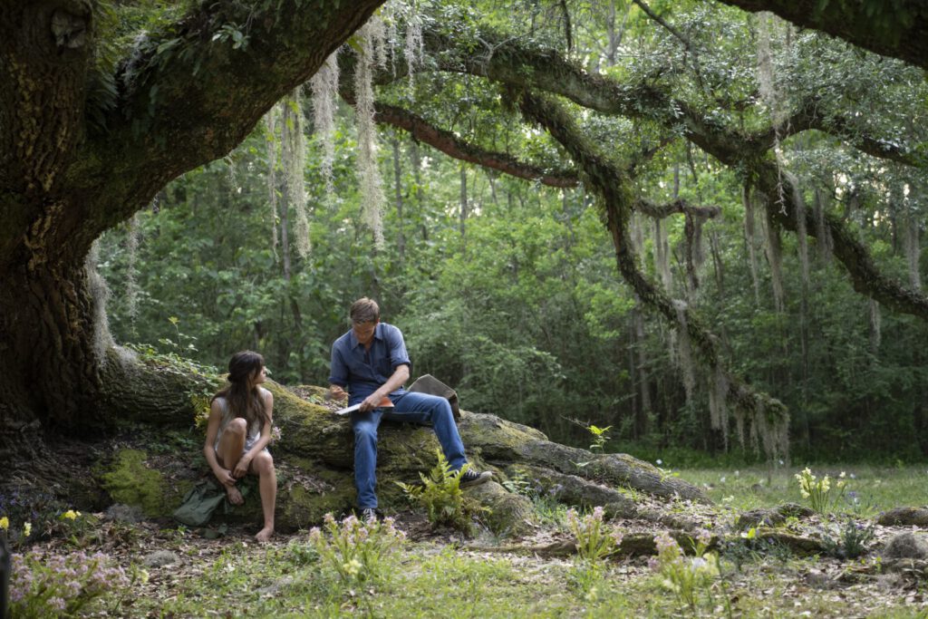 Bildauszug aus dem Film: Gesang der Flusskrebse. Die Hauptdarsteller sitzen auf den Wurzeln eines großen alten Baumes und üben lesen.